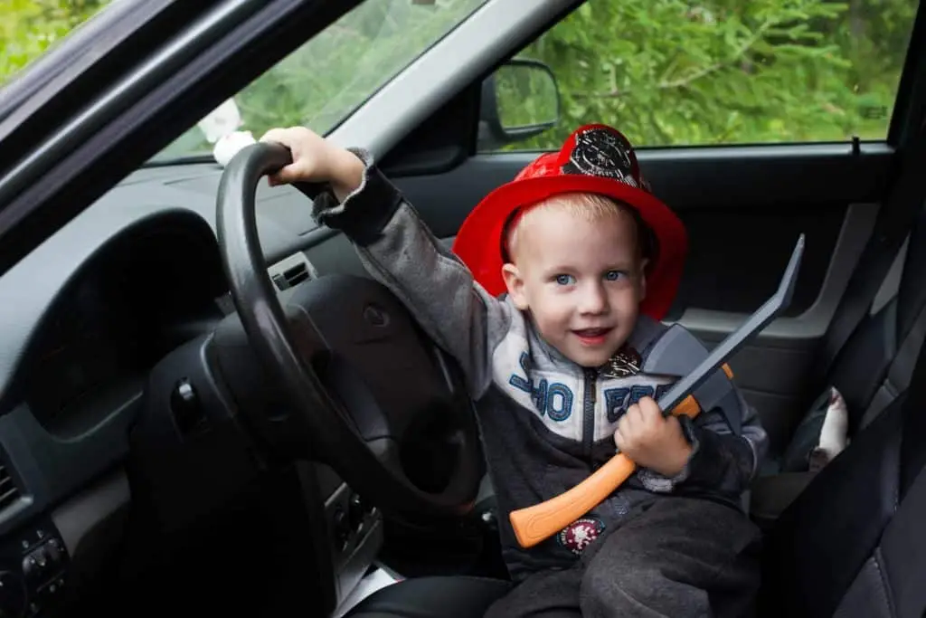 A young child with a firefighter's helmet and a toy axe playfully takes the driver's seat of a car, pretending to be a firefighter on the go.