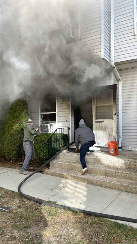 Two people with a hose direct water into a smoke-filled building. Dense smoke billows from the doorway and window. One person crouches on the stairs, while the other holds the hose. A red bucket is near the steps.