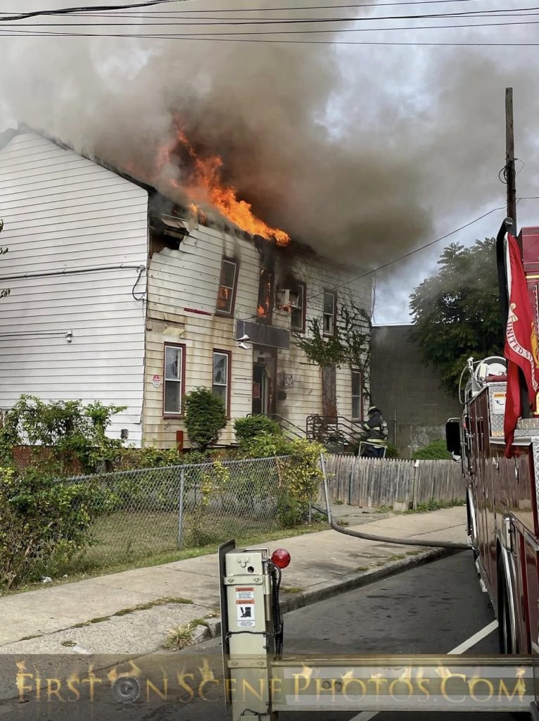 A two-story house is on fire, with flames and heavy smoke pouring from the upper windows. A fire truck is parked nearby, and a firefighter in gear stands close to the building, preparing to tackle the blaze.