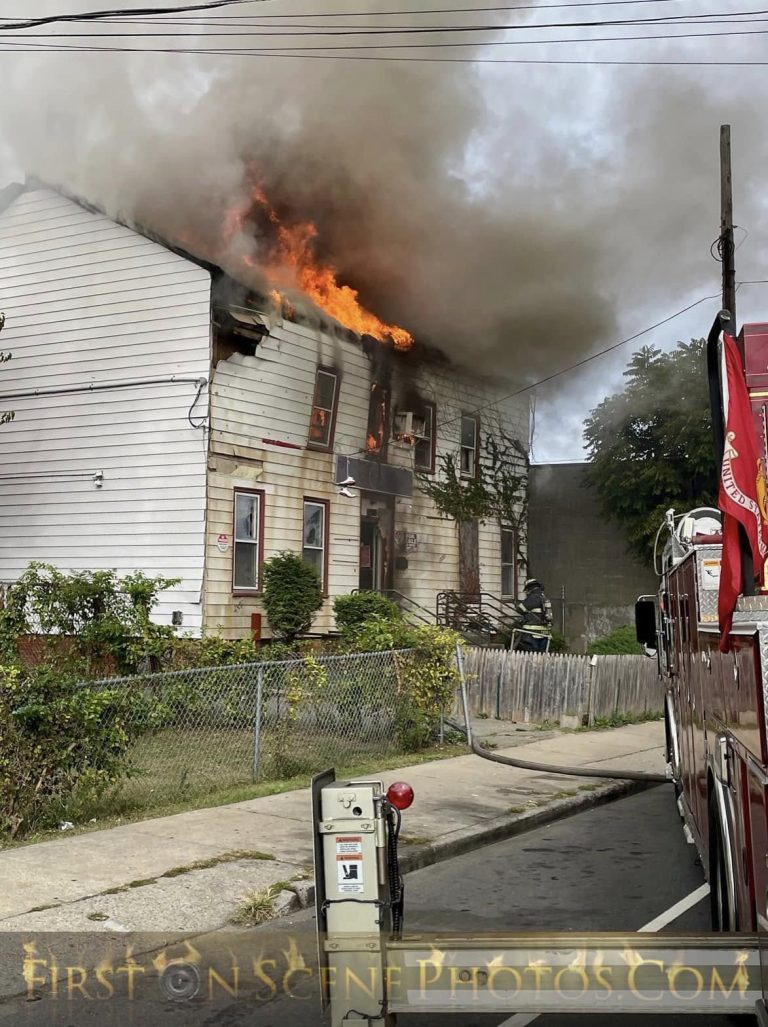A two-story house is on fire, with flames and heavy smoke pouring from the upper windows. A fire truck is parked nearby, and a firefighter in gear stands close to the building, preparing to tackle the blaze.