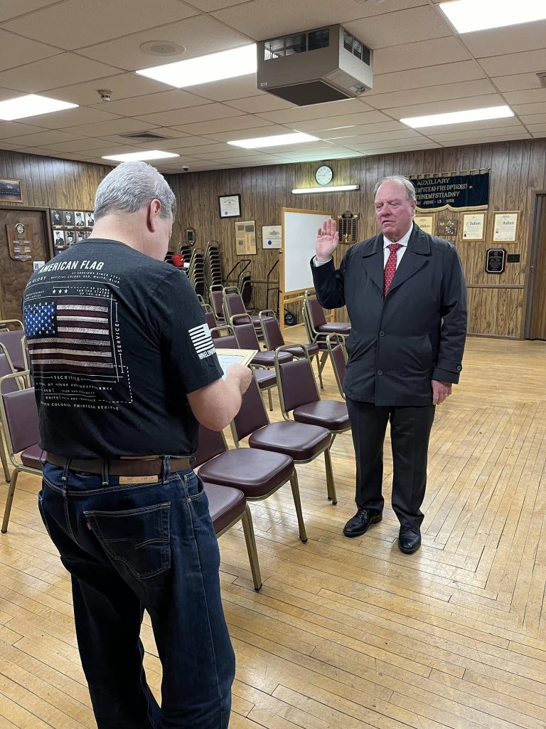 A man stands facing another man in a room with wood-paneled walls and rows of chairs. The man on the right is raising his right hand as if taking an oath, while the man on the left is taking a photo.