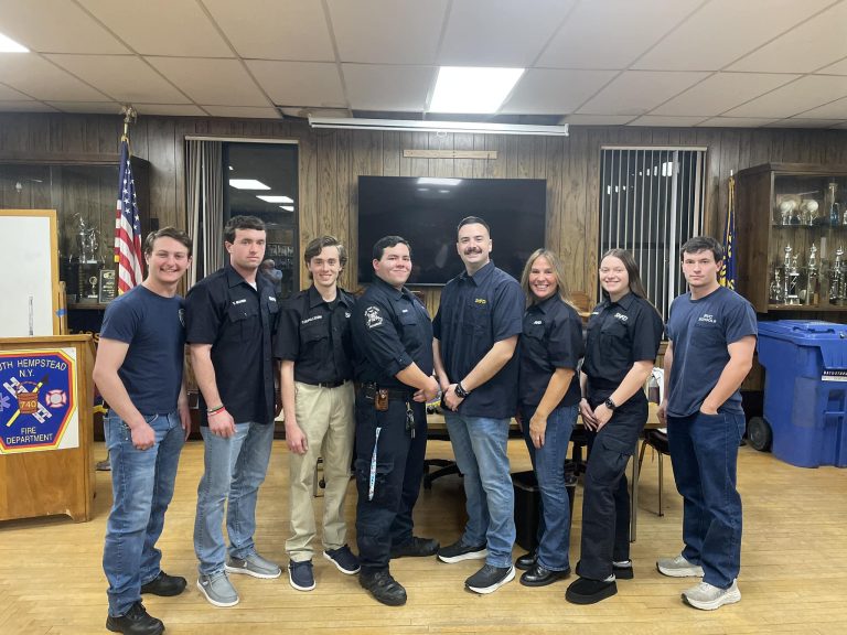 A group of eight people, five men and three women, stand smiling in a room with wood-paneled walls and shelves of trophies. They wear matching dark shirts. An American flag and a banner are visible in the background.
