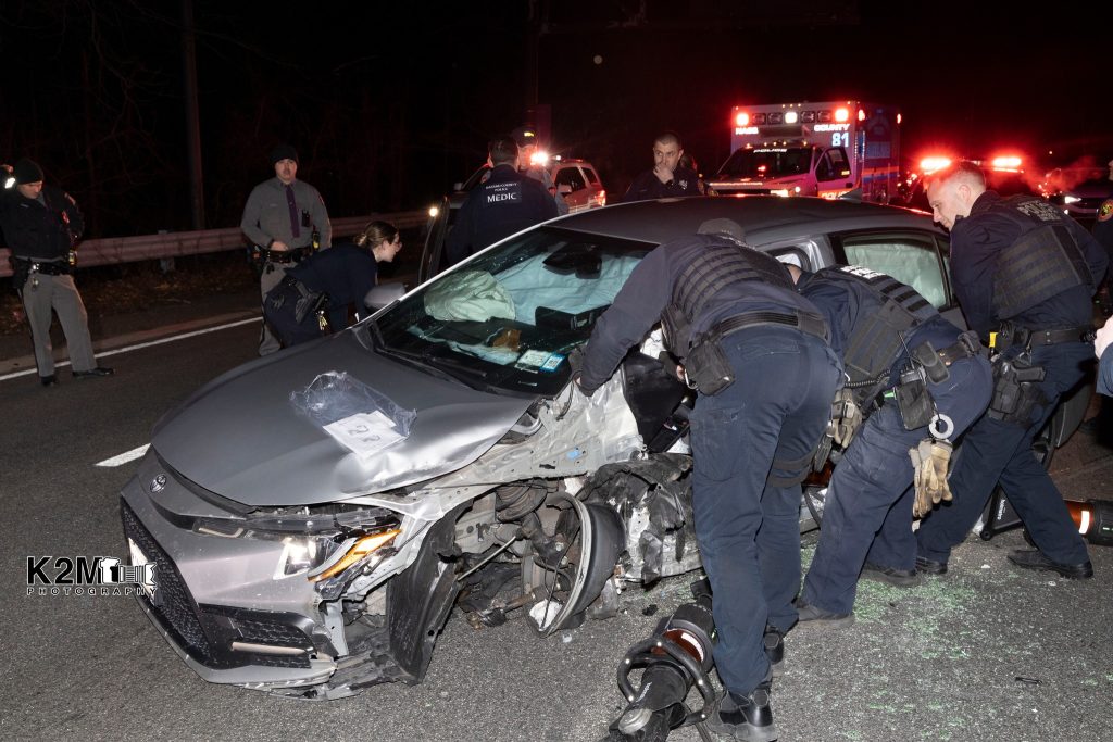 Police officers examine a damaged silver car involved in an accident on a dark road. The car's front and side are heavily dented, and debris is scattered. An ambulance with flashing lights is visible in the background, with officers standing nearby.