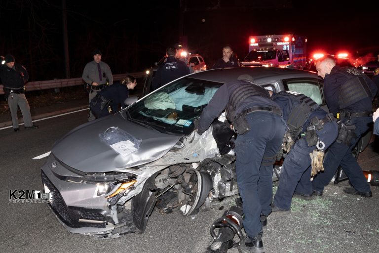 Police officers examine a damaged silver car involved in an accident on a dark road. The car's front and side are heavily dented, and debris is scattered. An ambulance with flashing lights is visible in the background, with officers standing nearby.