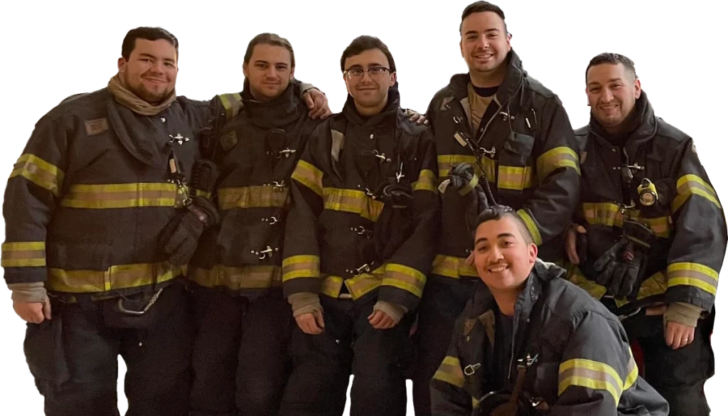 Six firefighters in full gear, smiling for a group photo. Five of them are standing, and one is kneeling in front. They all wear black uniforms with reflective yellow stripes.