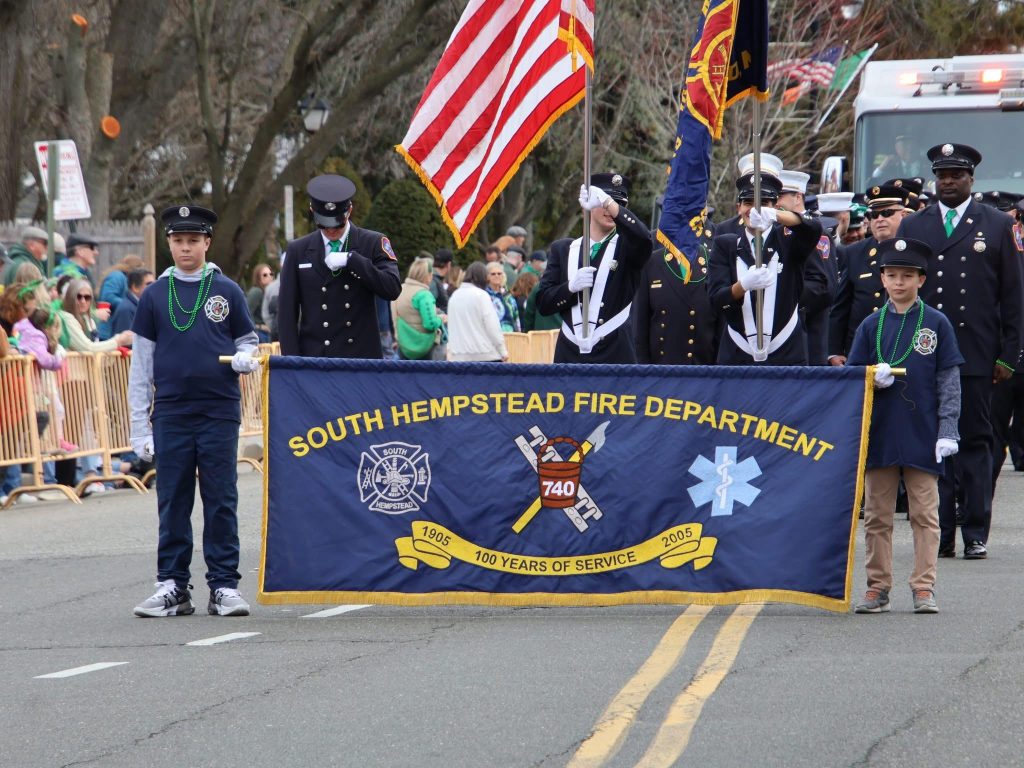 A parade scene with people from the South Hempstead Fire Department holding a large blue banner. They are marching on a street, accompanied by firefighters in uniform, with spectators in the background. An American flag is visible.