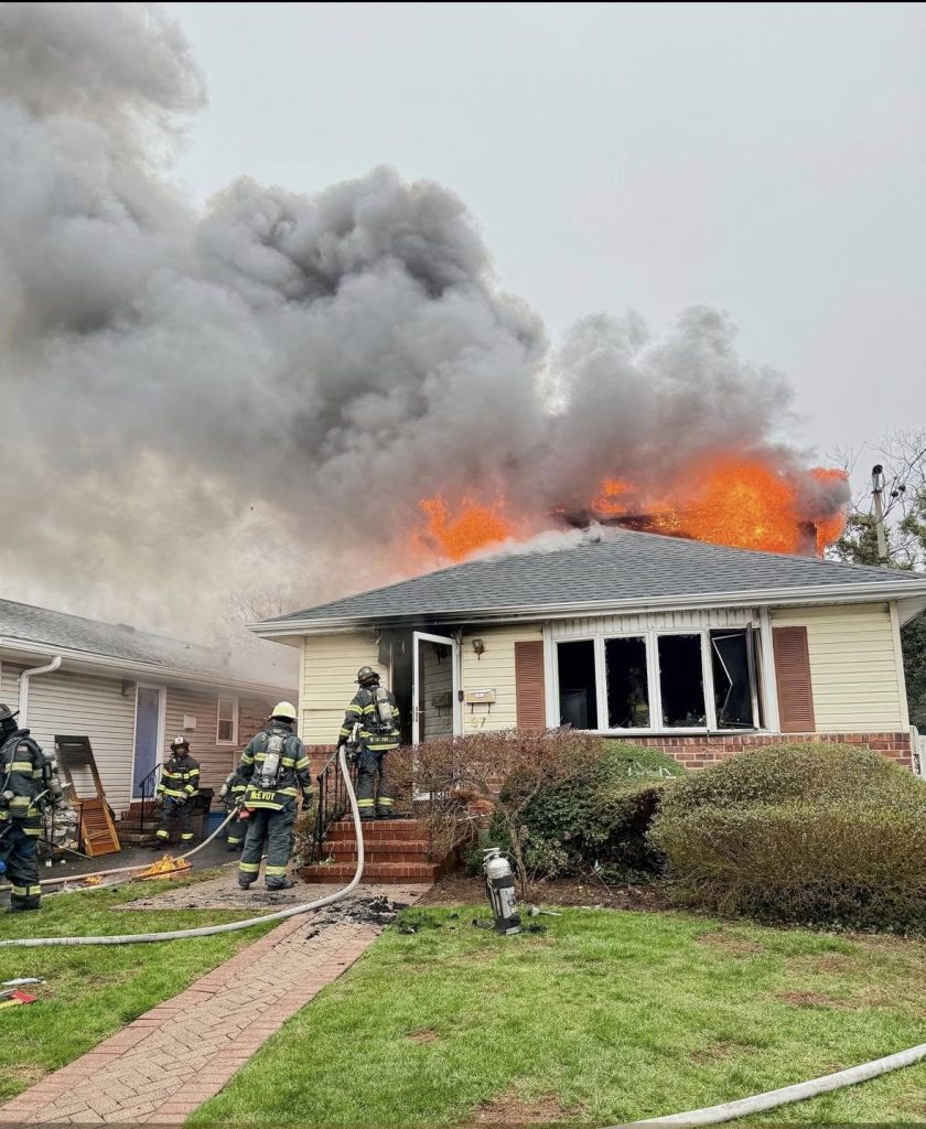 Firefighters battle a blaze at a single-story house with heavy smoke and flames erupting from the roof. Multiple hoses are in use, and the area is surrounded by green grass and bushes. The sky is overcast.