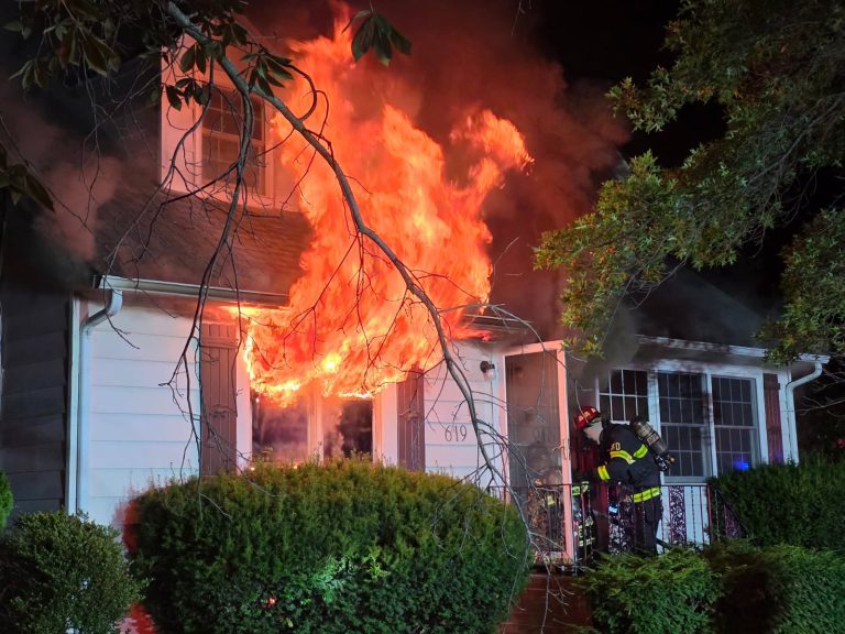 A house is engulfed in flames at night, with large fire coming from an upstairs window. Two firefighters in gear stand on the porch, preparing to enter. The scene is surrounded by bushes and tree branches in the foreground.